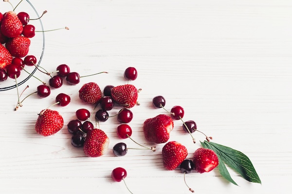 fresh cherries and strawberries scattered from bowl on white rustic wooden background. ripe juicy red  berries on table